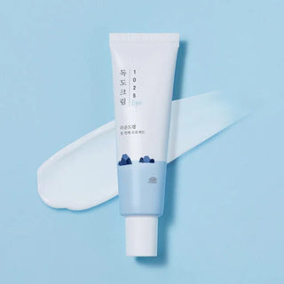 Tube ROUND LAB 1025 Dokdo Eye Cream à étiquette coréenne et swatch de crème blanche sur fond bleu ciel