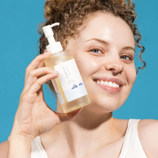 Femme souriante aux cheveux boucles tenant le flacon ROUND LAB 1025 Dokdo Cleansing Oil pres du visage