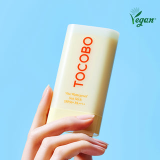 Main tenant le stick solaire TOCOBO Vita Waterproof SPF50+ avec badge Vegan sur fond bleu