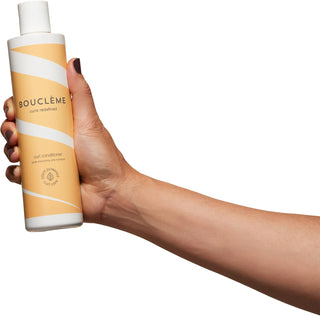 Main tenant le flacon Boucleme Curl Conditioner 300 ml orange et blanc sur fond blanc