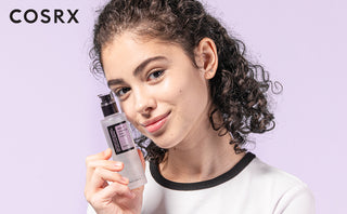 Jeune femme aux cheveux boucles tenant le flacon COSRX AHA 7 Whitehead Power Liquid sur fond mauve