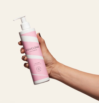 Main tenant le flacon pompe Boucleme Curl Cream rose et blanc sur fond beige