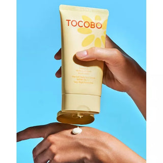 Mains appliquant une noisette de lotion solaire TOCOBO Vita Soft sur le dos de la main, fond bleu ciel