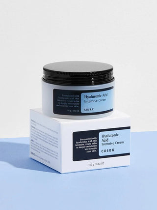 Pot COSRX Hyaluronic Acid Intensive Cream posé sur sa boîte blanche et bleue, décor bleu clair