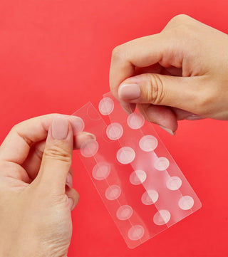 Deux mains tenant une planche de patchs hydrocolloïdes transparents COSRX sur fond rouge
