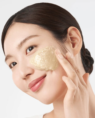 Femme aux cheveux bruns attachés appliquant le nettoyant exfoliant à l'avoine PURITO SEOUL sur la joue