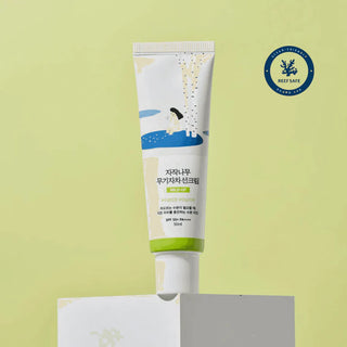 Round Lab Birch Mild-Up Sunscreen pose sur socle blanc avec badge Reef Safe, fond vert anis