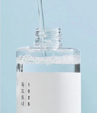 Round Lab 1025 Dokdo Toner - gros plan sur le goulot ouvert avec filet de liquide transparent sur fond bleu ciel