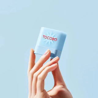 Main tenant le mini stick solaire TOCOBO bleu clair au logo soleil sur fond bleu pastel