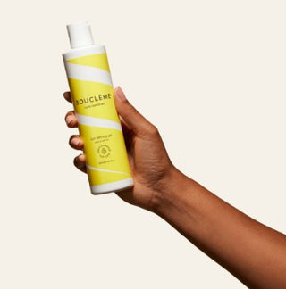 Main tenant le flacon Boucleme Curl Defining Gel jaune et blanc sur fond beige clair