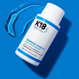 K18 Damage Shield flacon blanc 250ml pose a plat avec trace de gel bleu sur fond bleu vif