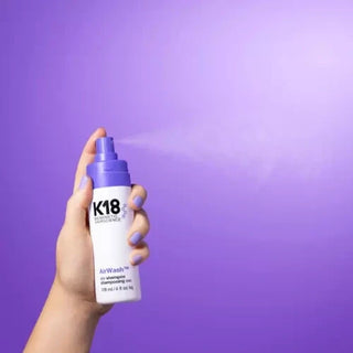 K18 AirWash Dry Shampoo - main tenant le flacon avec brume vaporisee sur fond violet