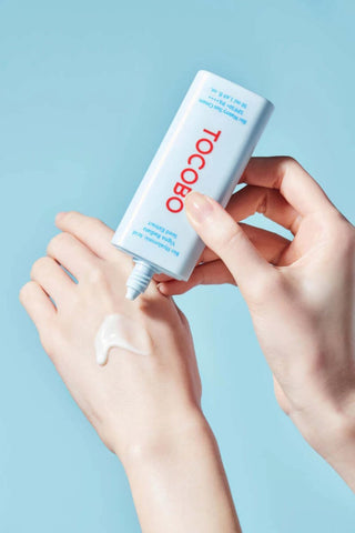 TOCOBO Bio Watery Sun Cream pressee sur le dos de la main, texture blanche fluide sur fond bleu