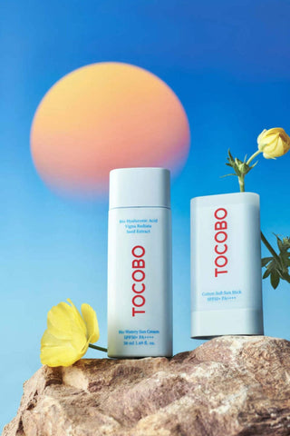 TOCOBO Bio Watery Sun Cream et Cotton Soft Sun Stick poses sur une roche avec fleurs et coucher de soleil