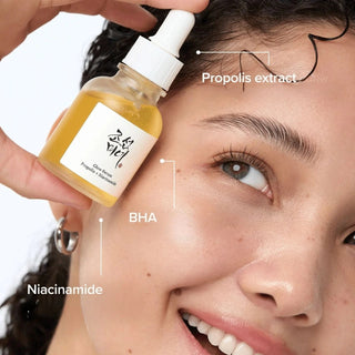 Femme souriante tenant le Glow Serum près de son visage - actifs propolis, BHA et niacinamide annotés