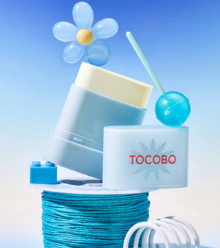 Composition ludique avec le stick solaire TOCOBO ouvert, sucette bleue et fleur - texture crème visible